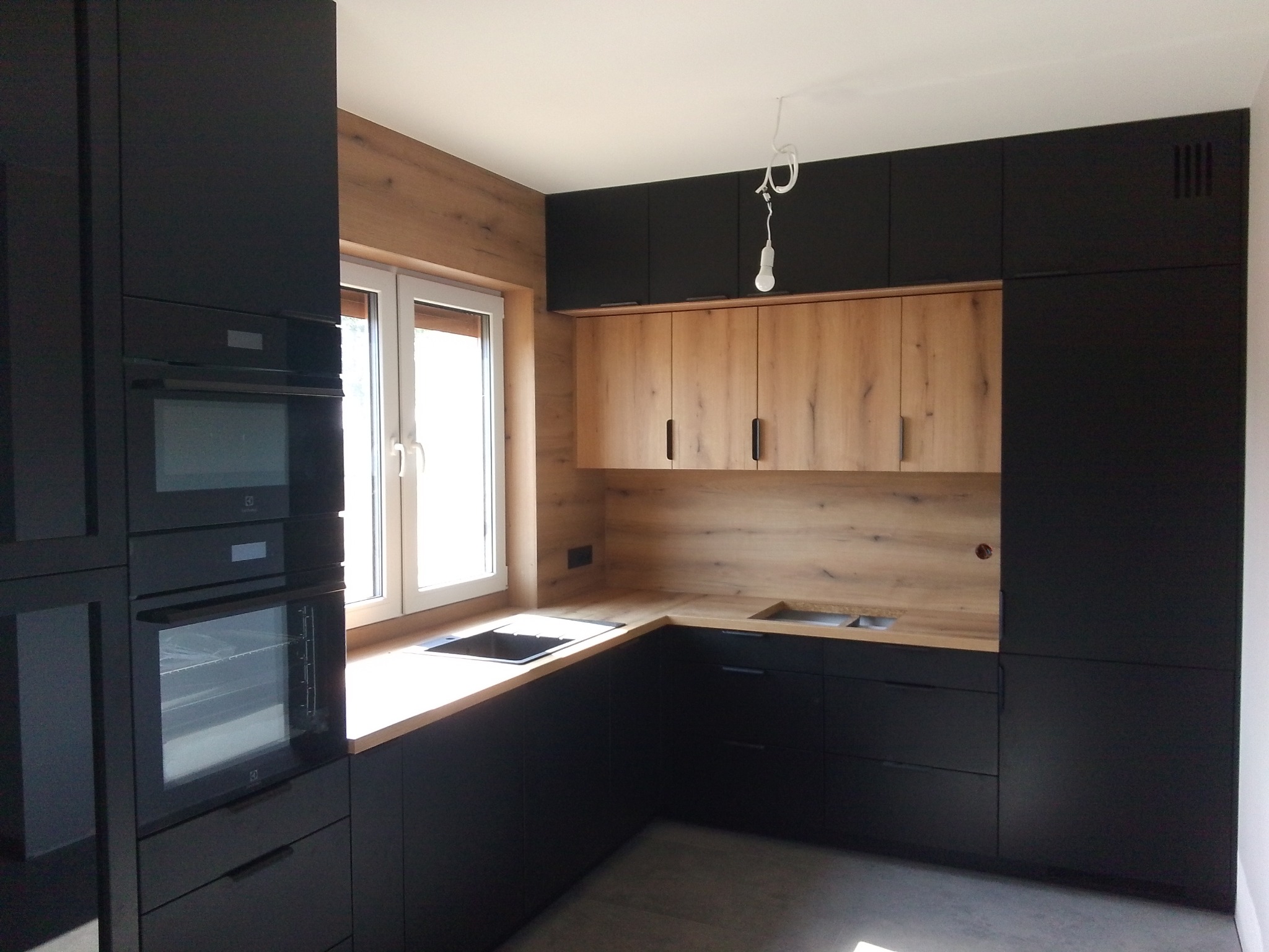 Nowoczesna, narożna kuchnia z czarnymi frontami i drewnopodobnym blatem. Widoczne okno z jasnym światłem i wisząca żarówka. Minimalistyczny design.
