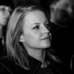 Portret kobiety w czerni i bieli, delikatny uśmiech, profesjonalny wygląd, w tle zamazane postacie.