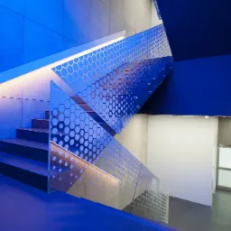 Nowoczesne schody z dekoracyjną, szklaną balustradą z motywem plastra miodu, oświetlone niebieskim światłem. Widok z dołu, betonowe ściany i schody.
