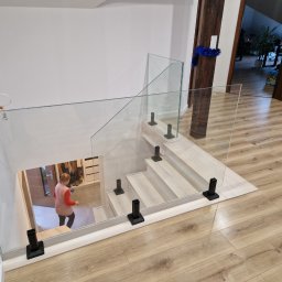 F.U.H.P. Szkło-Plast Curyło - Szklana balustrada na schodach z czarnymi mocowaniami, widok z góry, osoba w oddali. Jasne drewno schodów i podłogi, białe ściany. Nowoczesny design.
