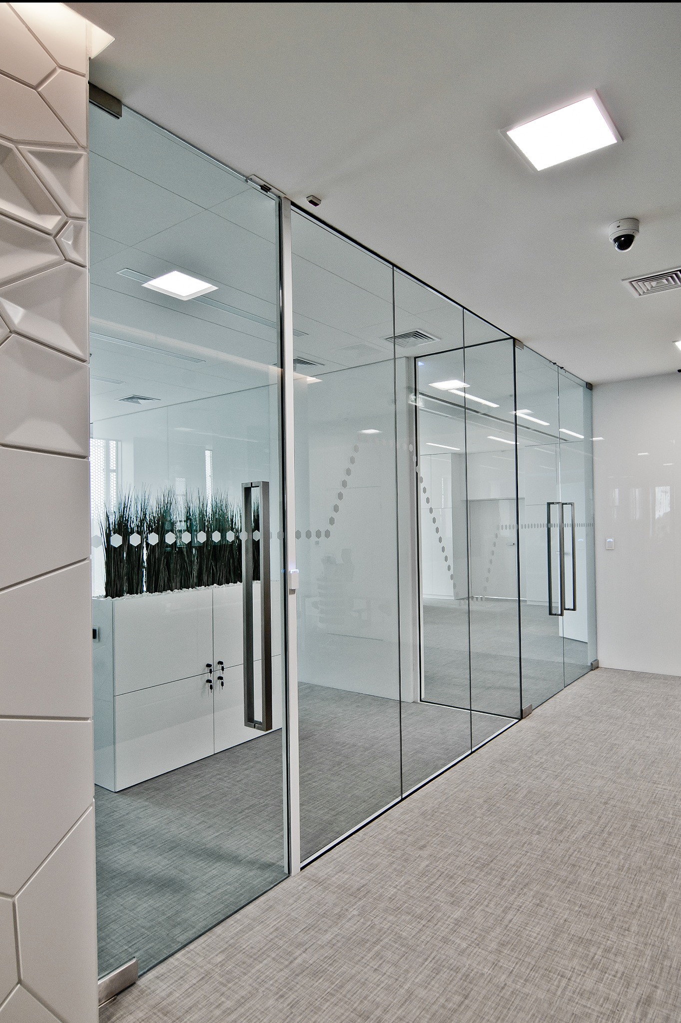 Szklane drzwi przesuwne w biurze z minimalistycznym designem, z widoczną dekoracją w postaci roślin w białej donicy. Wykończenie w odcieniach bieli i szarości.