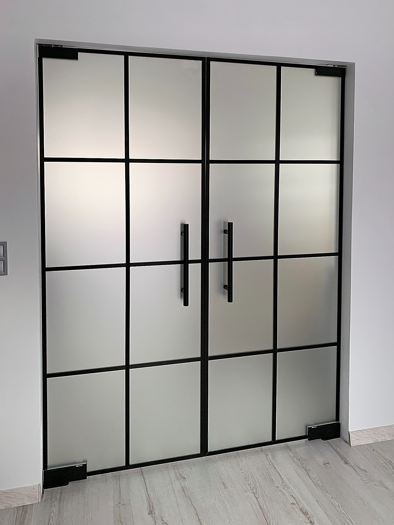 Dwuskrzydłowe drzwi szklane, matowe, z czarnymi szprosami i uchwytami, widoczne zawiasy podłogowe. Minimalistyczny design, jasne wnętrze.