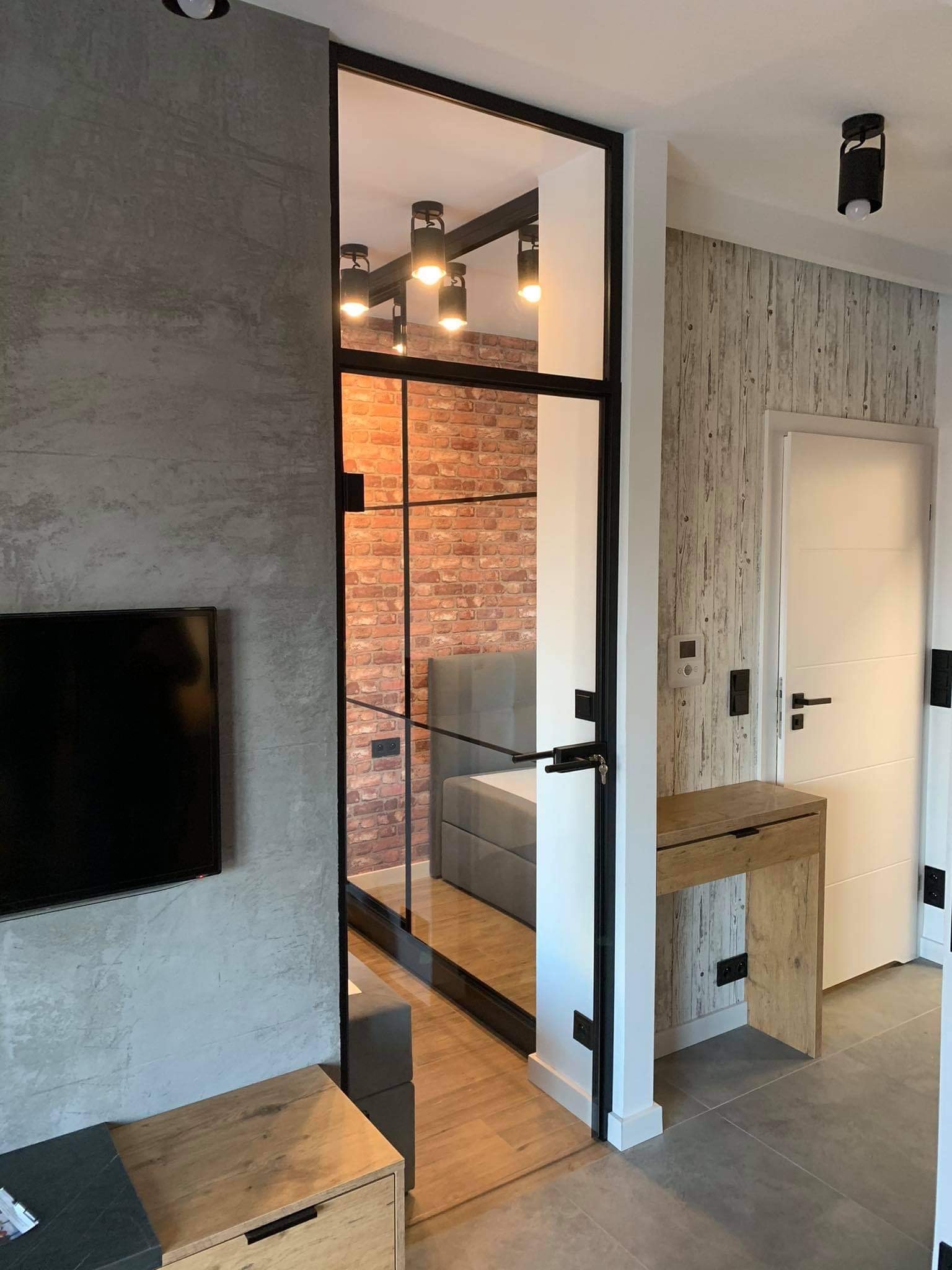 Nowoczesne drzwi z czarną ramą i szybą, oddzielające pokój z ceglaną ścianą. Widoczny fragment szafki RTV i drzwi wejściowe.