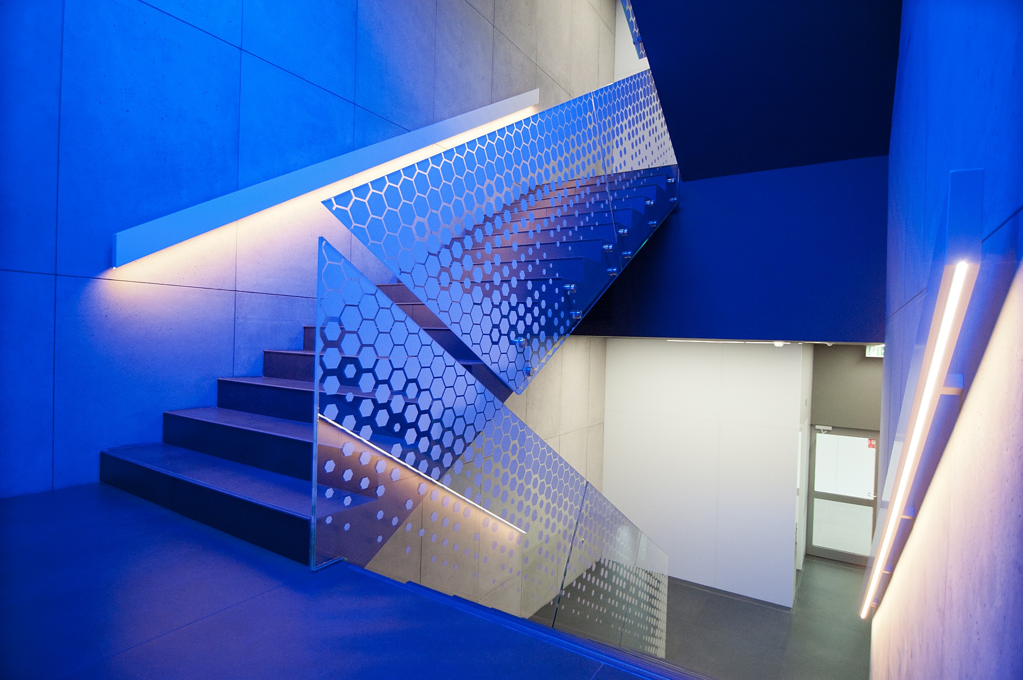 Nowoczesne schody z dekoracyjną, szklaną balustradą z motywem plastra miodu, oświetlone niebieskim światłem. Widok z dołu, betonowe ściany i schody.