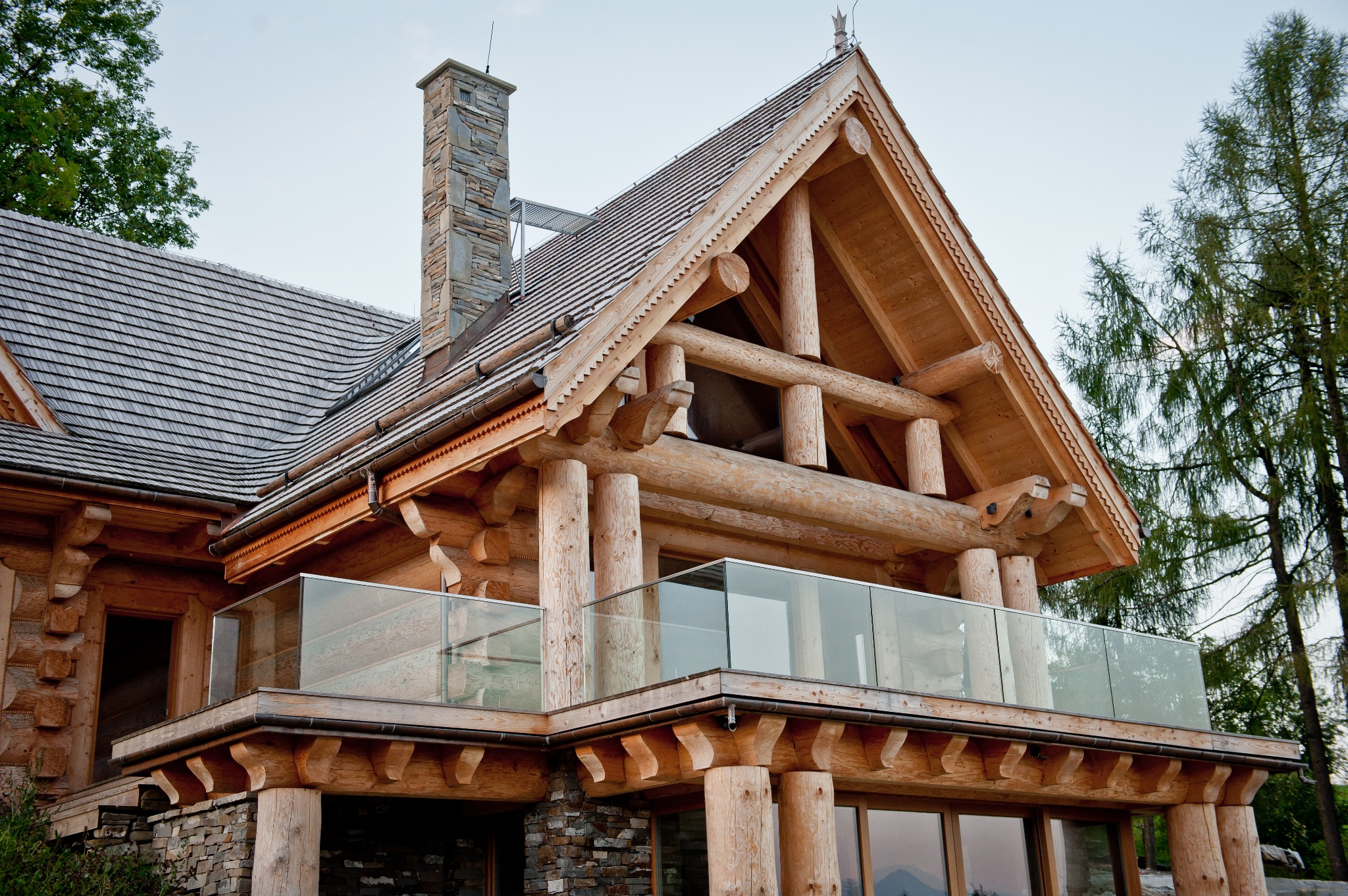 Szklana balustrada balkonu w drewnianym domu z bali, widok z dołu, na tle drzew i nieba. Balustrada z przezroczystego szkła, mocowana do drewnianej konstrukcji.