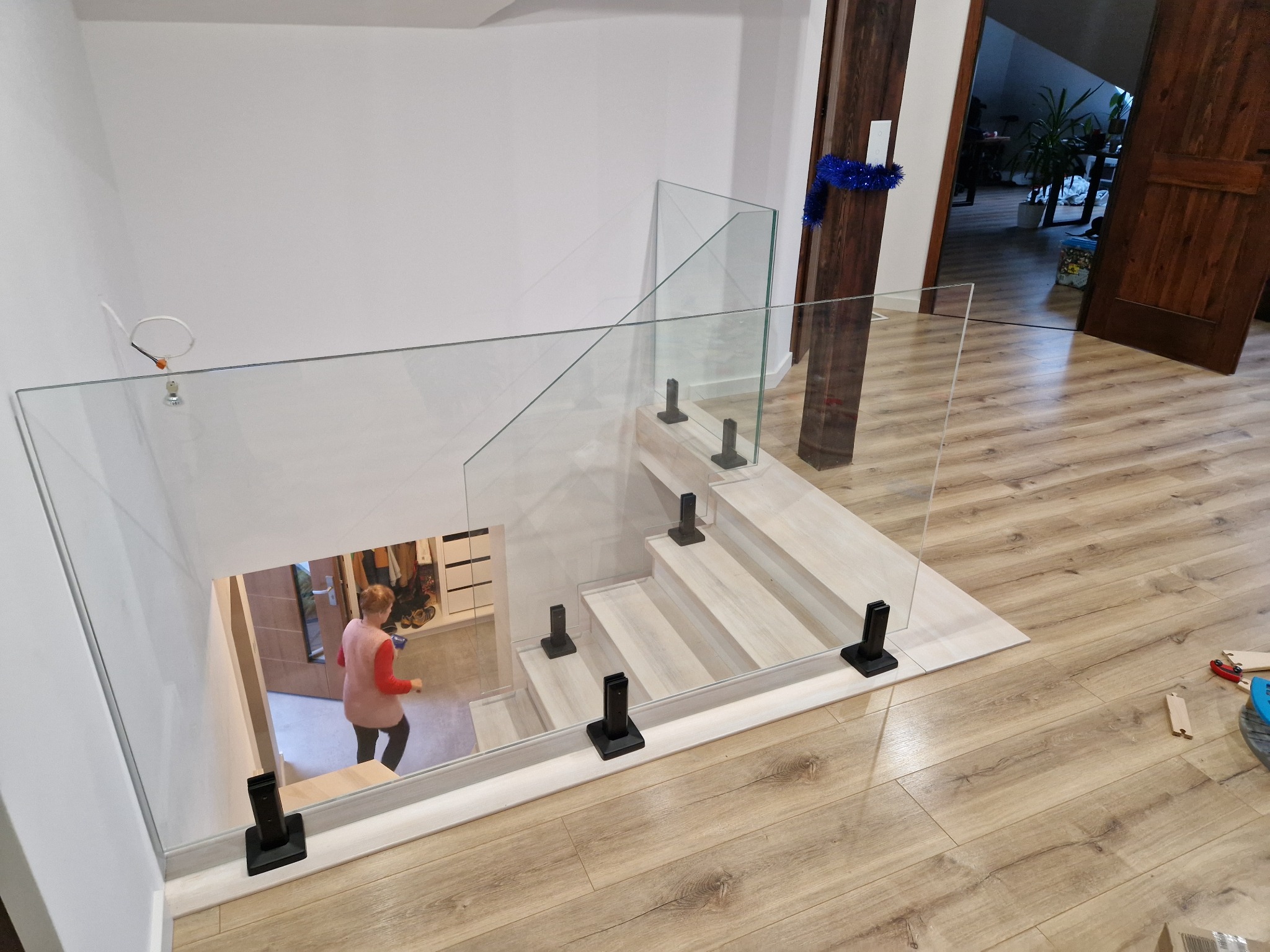 Szklana balustrada na schodach z czarnymi mocowaniami, widok z góry, osoba w oddali. Jasne drewno schodów i podłogi, białe ściany. Nowoczesny design.