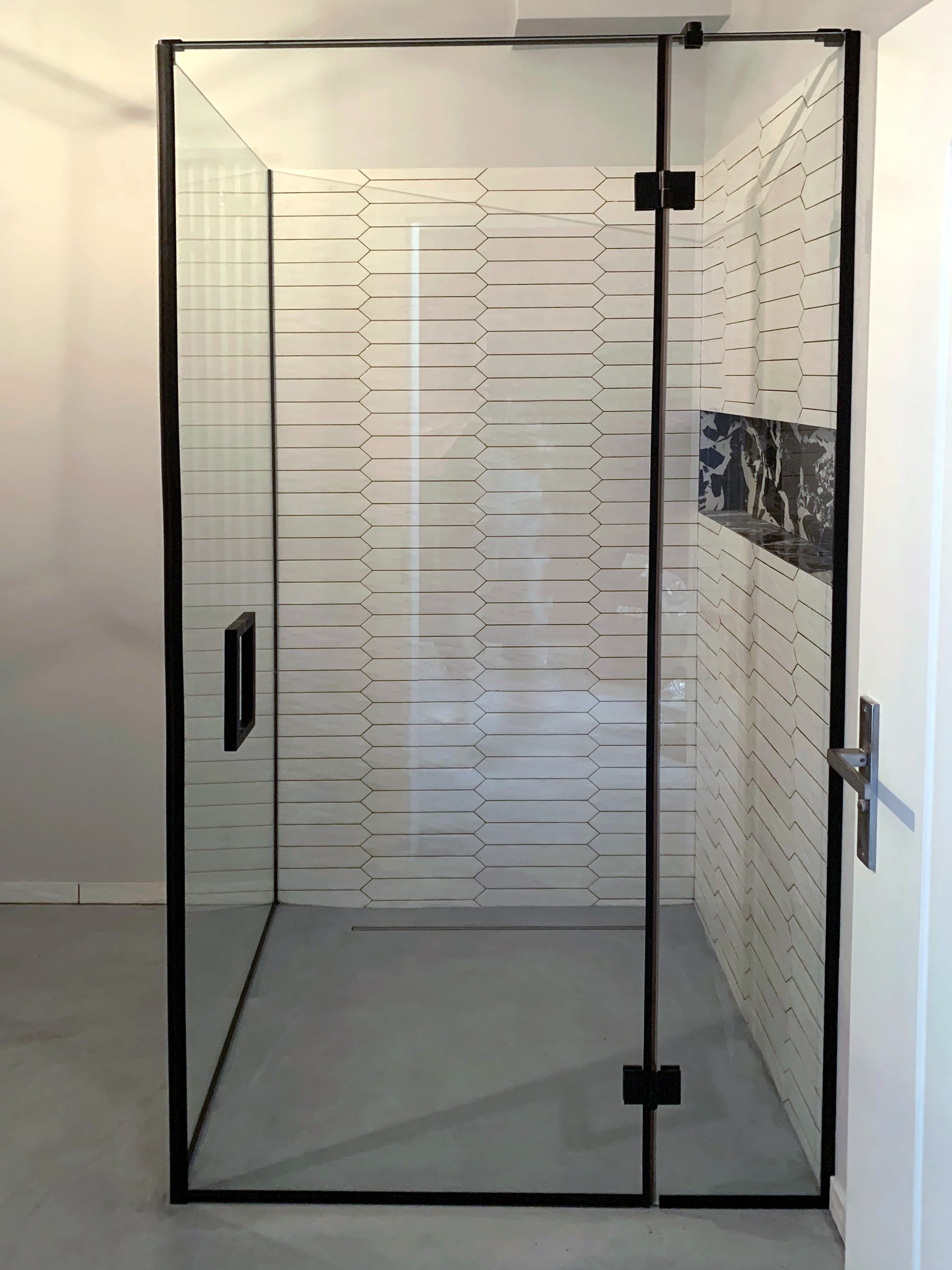 Nowoczesna kabina prysznicowa ze szklaną ścianą i czarnymi profilami na tle białych płytek w kształcie rombów. Minimalistyczny design.
