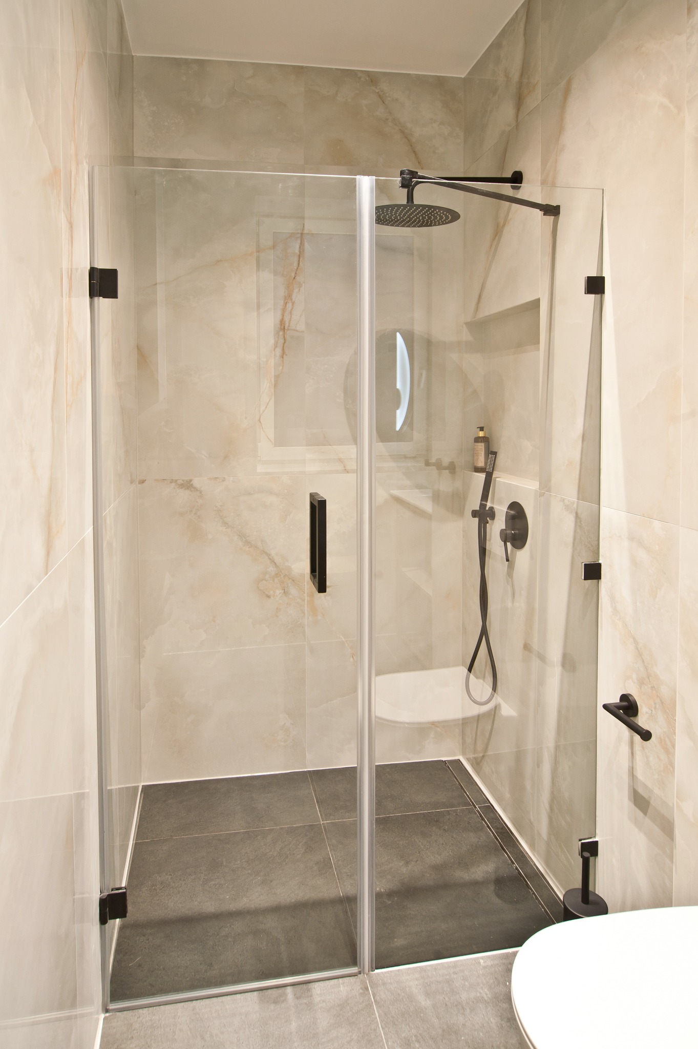 Kabina prysznicowa ze szklanymi drzwiami, czarne okucia, beżowe płytki imitujące marmur, szara podłoga. Widoczne bateria i deszczownica w kolorze czarnym.