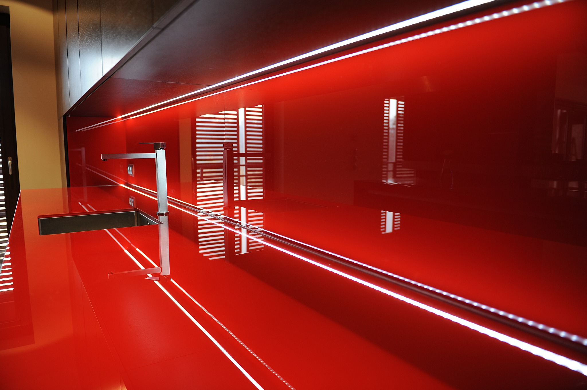 Błyszcząca, czerwona szklana ściana w kuchni z podświetleniem LED. Nowoczesny zlew i bateria odbijają światło, tworząc geometryczne wzory.