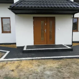 Nowoczesny dom z ciemną kostką brukową obramowaną białym pasem, prowadzącą do drewnianych drzwi wejściowych; widoczne okna z roletami i czarny dach.