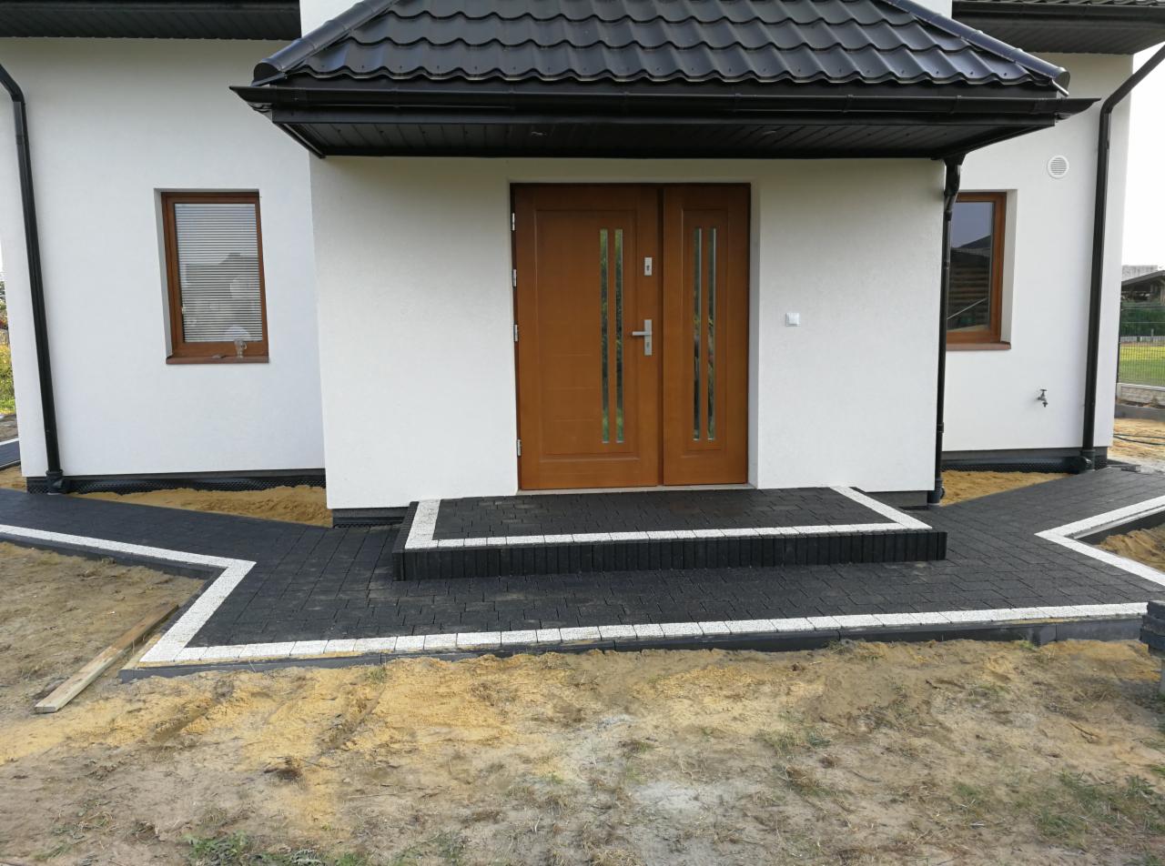 Nowoczesny dom z ciemną kostką brukową obramowaną białym pasem, prowadzącą do drewnianych drzwi wejściowych; widoczne okna z roletami i czarny dach.