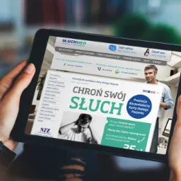 Osoba trzyma tablet prezentujący stronę internetową firmy Słuchmed z informacją o promocji dla posiadaczy Karty Stałego Pacjenta, skupiającą się na ochronie słuchu.