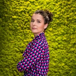 Portret kobiety z kręconymi włosami w koszuli z geometrycznym wzorem na tle ściany z mchu.