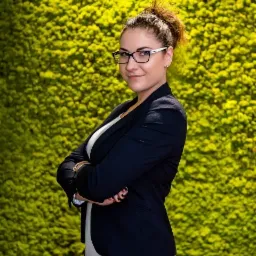 Portret kobiety w okularach, czarnej marynarce i wzorzystych spodniach, stojącej na tle ściany z mchu.