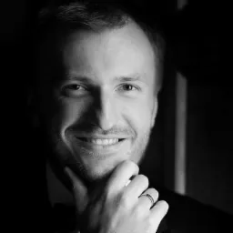 Czarno-biały portret uśmiechniętego mężczyzny w garniturze z brodą, trzymającego dłoń pod brodą, widoczny pierścionek na palcu.