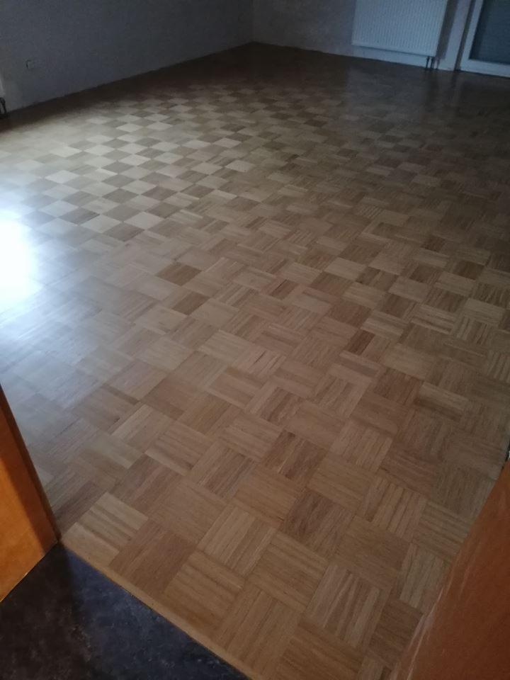 Parkiet ułożony w szachownicę z małych, drewnianych kostek, widok z progu pokoju, ściany pomalowane na szaro, w tle grzejnik.