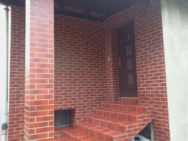 Wykończone wejście do budynku z brązowymi płytkami imitującymi cegłę, obejmujące schody wejściowe i ściany, z ciemnymi fugami i widocznymi elementami instalacji zewnętrznej.