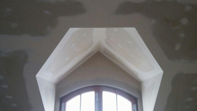 Nisko kątowe ujęcie fragmentu sufitu z oknem łukowym w trakcie prac wykończeniowych, widoczne ślady po szpachlowaniu na suficie i narożnikach okna.