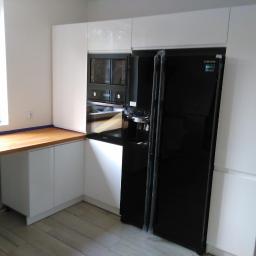 Meble Zuza - Biała zabudowa kuchenna z czarną lodówką Samsung i piekarnikiem w zabudowie, drewniany blat roboczy pod oknem, szare panele podłogowe.