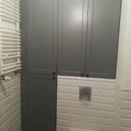 Szara szafa łazienkowa z trzema drzwiami, dopasowana do zabudowy toalety w białych, wypukłych płytkach.