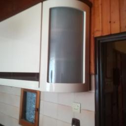 Meble Zuza - Biała szafka kuchenna z zaokrąglonym narożnikiem i matową szybą, zamontowana na ścianie z jasnych płytek, widoczny fragment drewnianej boazerii i gniazdko elektryczne.