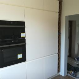 Meble Zuza - Biała zabudowa kuchenna z piekarnikiem i szafkami, wykończona brązową listwą, widoczna w trakcie montażu, z narzędziami w tle.