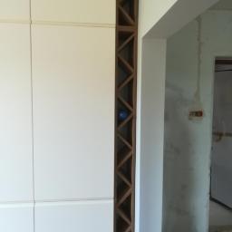 Meble Zuza - Biała szafa z drewnianymi półkami w kształcie rombów, widoczna butelka w jednej z przegródek, surowy stan wykończenia wnętrza w tle.