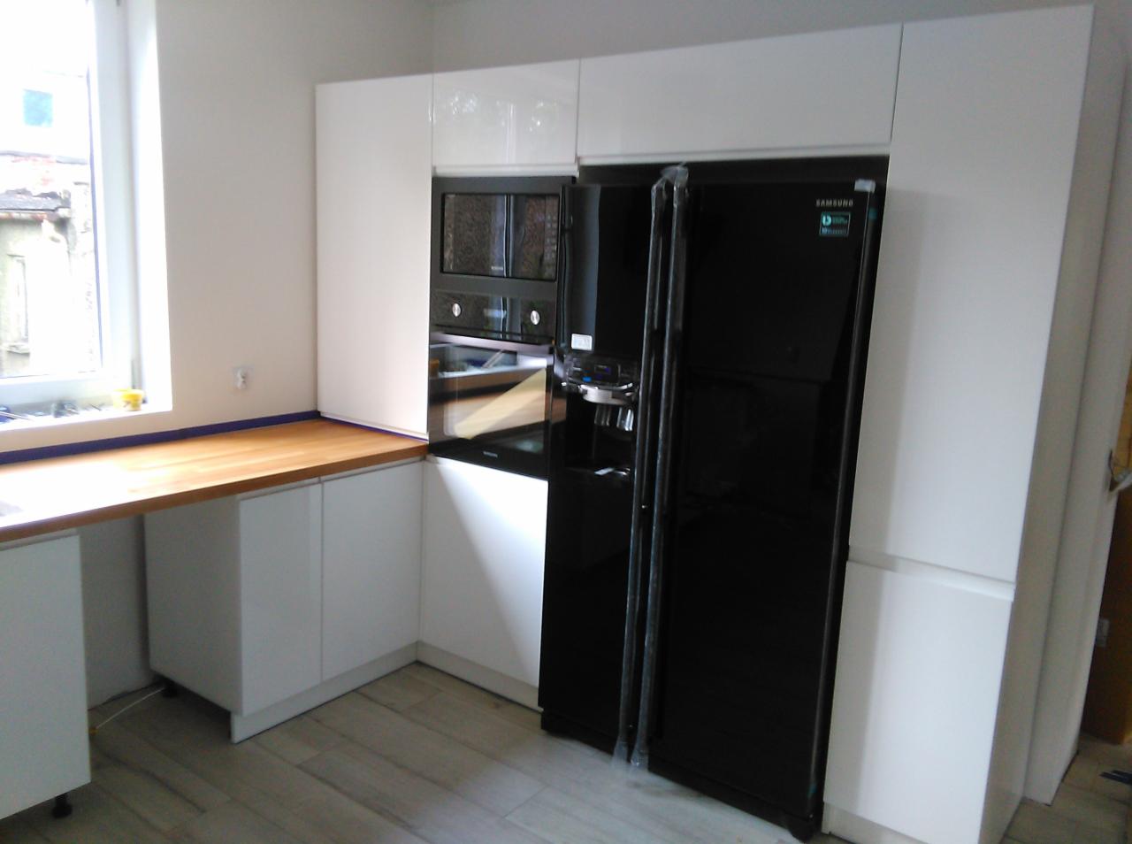 Biała zabudowa kuchenna z czarną lodówką Samsung i piekarnikiem w zabudowie, drewniany blat roboczy pod oknem, szare panele podłogowe.