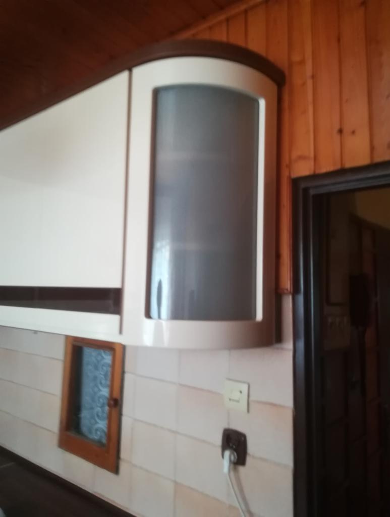 Biała szafka kuchenna z zaokrąglonym narożnikiem i matową szybą, zamontowana na ścianie z jasnych płytek, widoczny fragment drewnianej boazerii i gniazdko elektryczne.