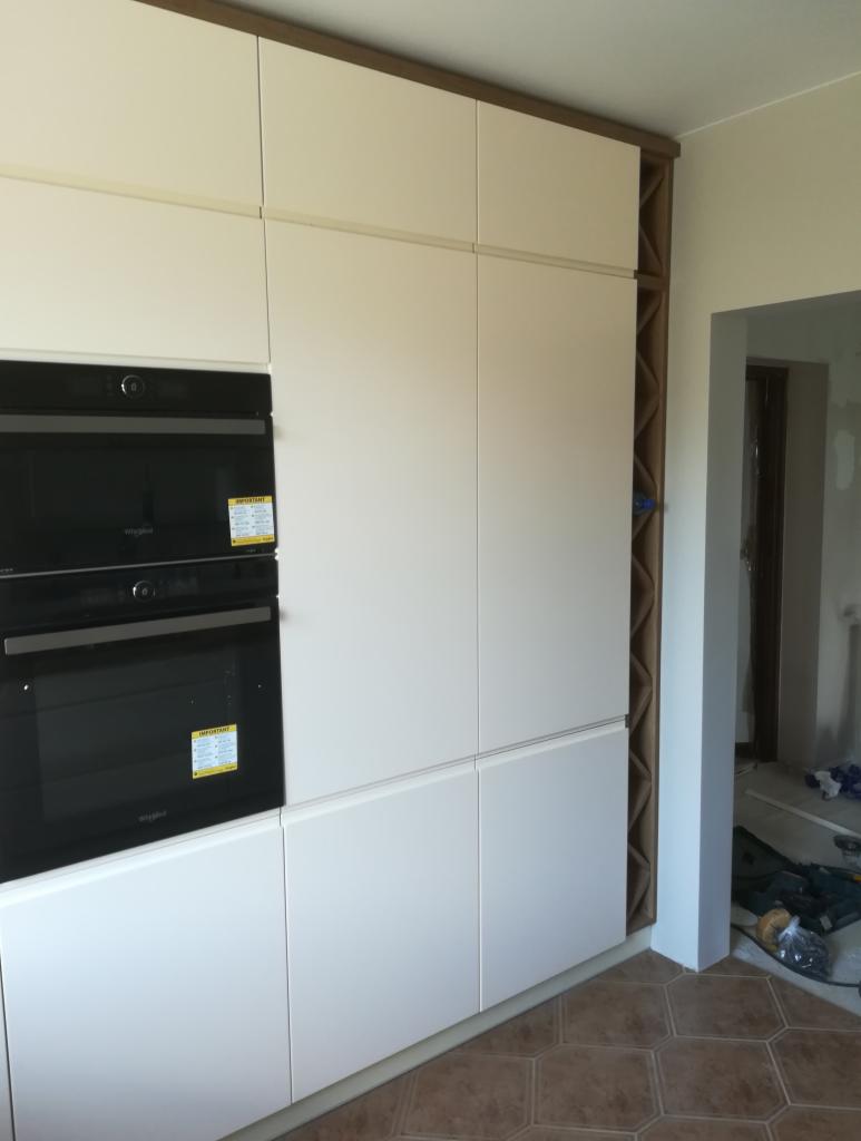 Biała zabudowa kuchenna z piekarnikiem i szafkami, wykończona brązową listwą, widoczna w trakcie montażu, z narzędziami w tle.