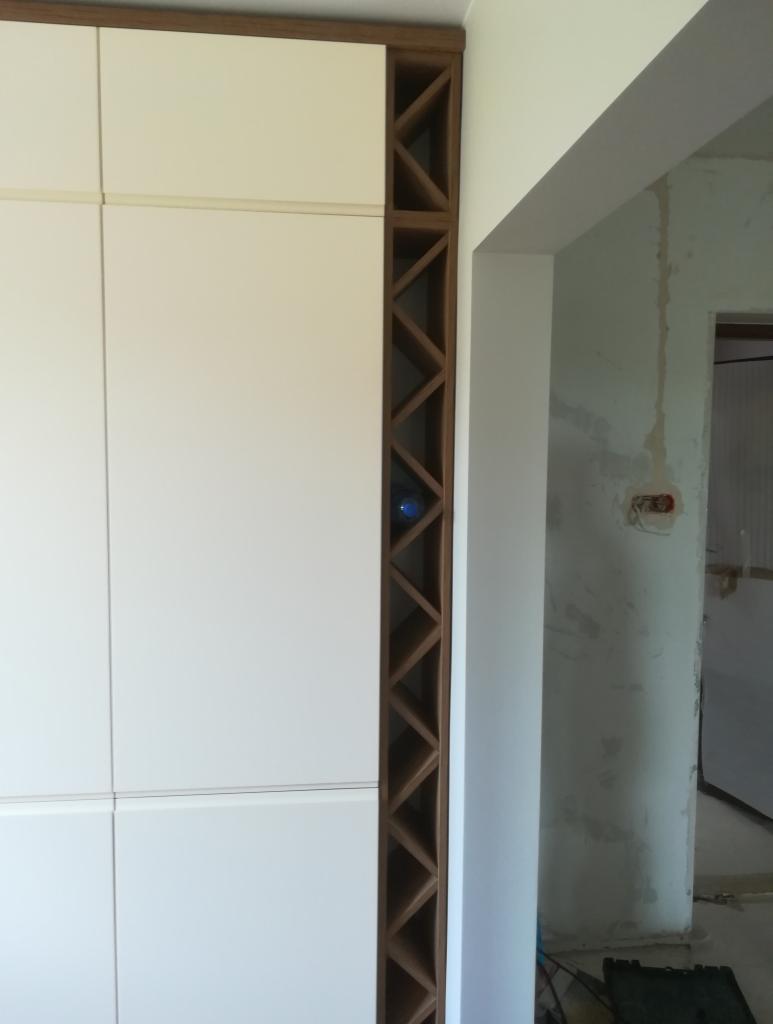 Biała szafa z drewnianymi półkami w kształcie rombów, widoczna butelka w jednej z przegródek, surowy stan wykończenia wnętrza w tle.