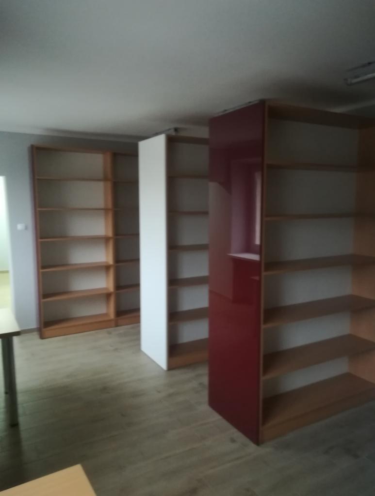 Wnętrze z regałami: drewniane z otwartymi półkami, biały moduł i czerwony, błyszczący element. Widoczne puste półki i podłoga imitująca drewno.