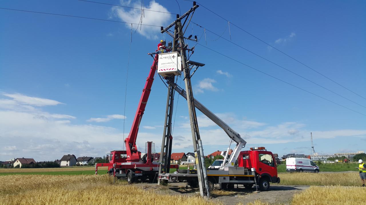 Prace na słupie wysokiego napięcia z użyciem dwóch podnośników koszowych, w tle pole i zabudowania mieszkalne.