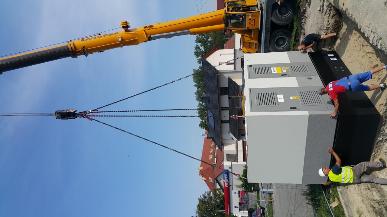 Montaż dużej szafy elektrycznej za pomocą dźwigu na placu budowy, widoczni dwaj pracownicy w strojach roboczych, jeden w kasku, drugi w czerwonym kombinezonie.