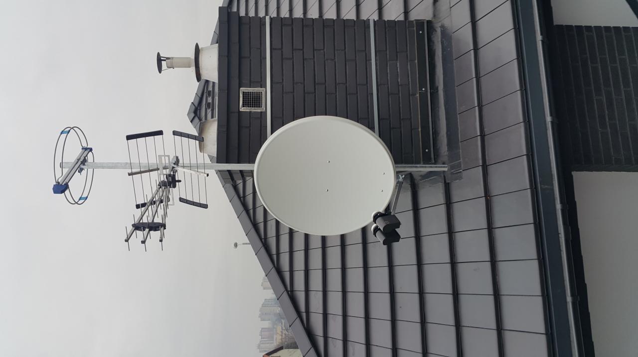 Anteny telewizyjne i satelitarne zamontowane na dachu budynku z ciemną dachówką, widok z dołu na tle pochmurnego nieba.