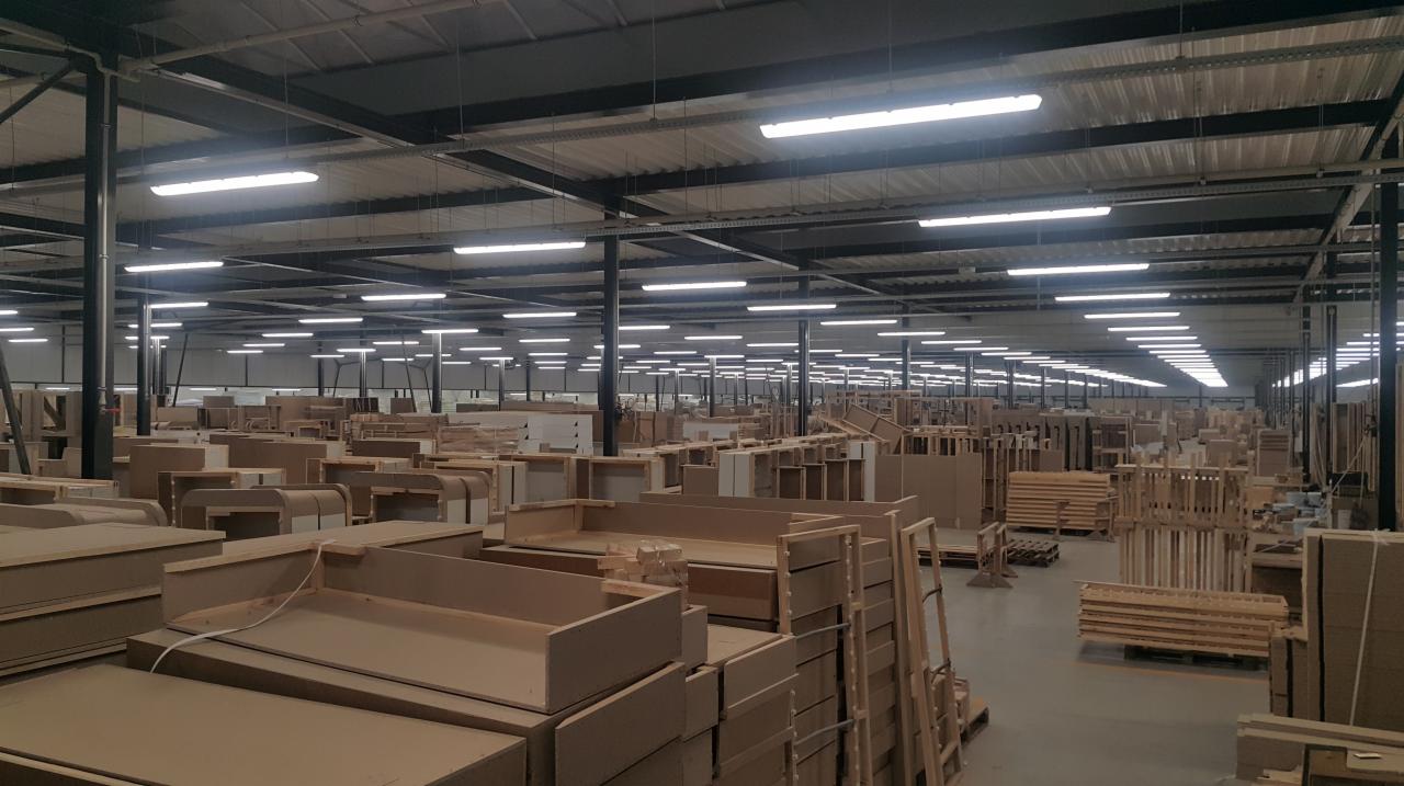 Rozległa hala produkcyjna oświetlona rzędami lamp, wypełniona stertami surowych, niezmontowanych elementów meblowych z płyty MDF i drewnianych palet, widok w głąb hali.