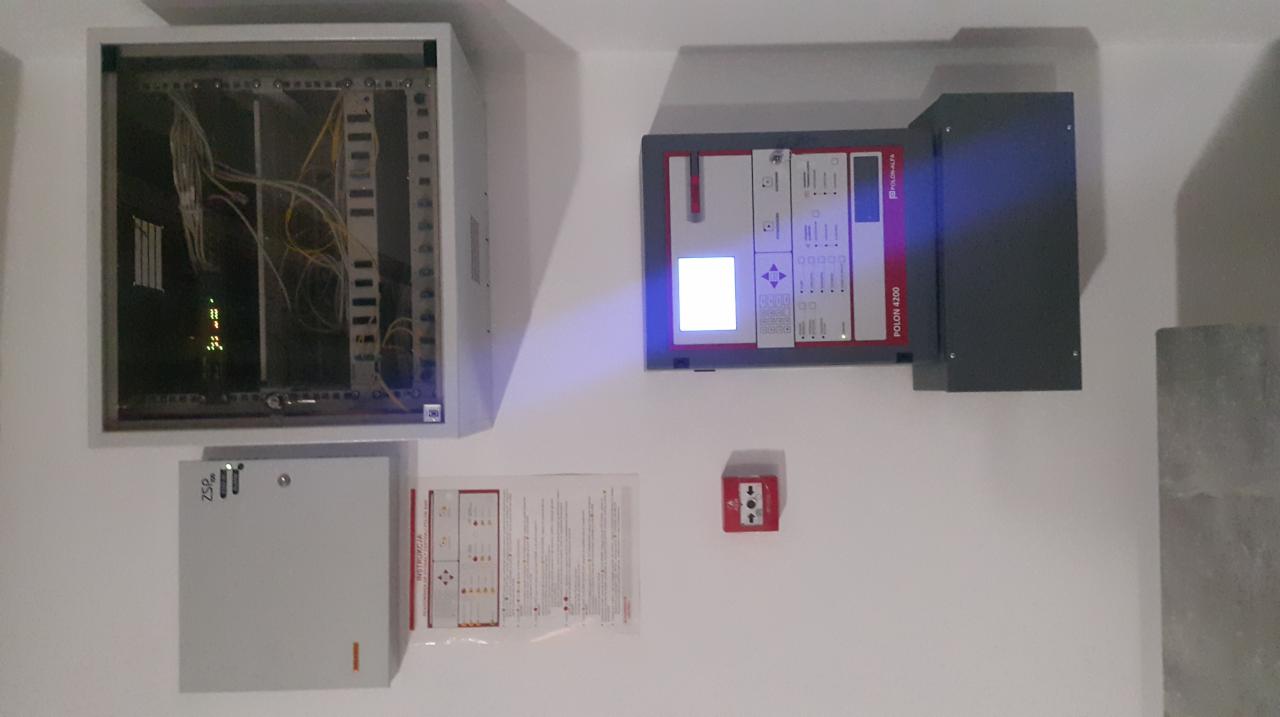 Panel sterowania systemem alarmowym Polon 4200 z instrukcją obsługi obok, centrala z okablowaniem w obudowie oraz przycisk alarmowy na białej ścianie.