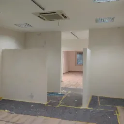 Wnętrze w trakcie prac wykończeniowych: świeżo pomalowane ściany w odcieniach bieli, zabezpieczona podłoga z widocznymi fragmentami paneli, oświetlenie sufitowe rastrowe i liniowe, widoczne...
