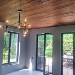 Jasne wnętrze z drewnianym sufitem, nowoczesnym żyrandolem kulistym i dużymi oknami wychodzącymi na zielony ogród.