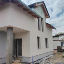 Nowo otynkowany budynek jednorodzinny z widocznymi elementami drewnianymi konstrukcji dachu i orynnowaniem, teren wokół w trakcie prac wykończeniowych, widoczne przewody i materiały budowlane.