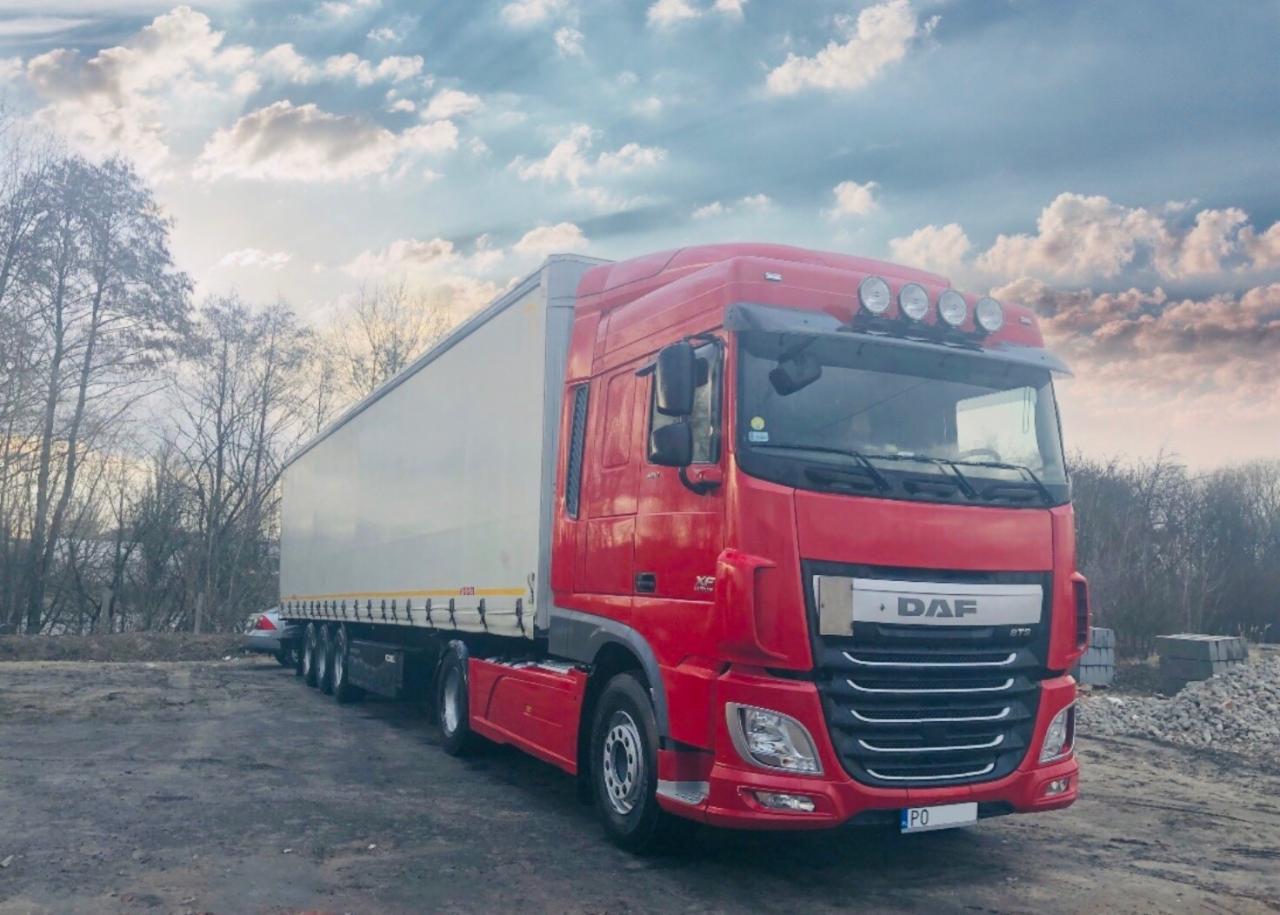 Czerwona ciężarówka DAF z białą naczepą zaparkowana na placu, widoczne drzewa w tle i pochmurne niebo.