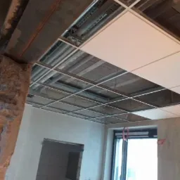 Montaż podwieszanego sufitu kasetonowego w pomieszczeniu z widoczną instalacją pod sufitem, częściowo wykończone ściany i otwór drzwiowy bez ościeżnicy.