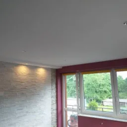 Pok&oacute;j po remoncie: ściana wyłożona dekoracyjnym kamieniem, świeżo pomalowane ściany w kolorze burgundowym i biały sufit z oświetleniem punktowym, okna z widokiem na zieleń.