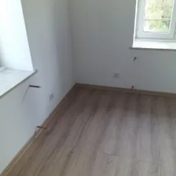 Pokój po remoncie: świeżo położone panele podłogowe w odcieniach brązu, białe ściany z wystającymi rurkami instalacji, okno z widokiem na zieleń.