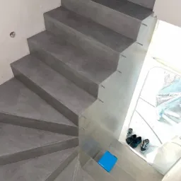 Betonowe schody spiralne z widocznymi metalowymi wspornikami, niebieskie pudełko i para butów na dolnym biegu, widok z góry.