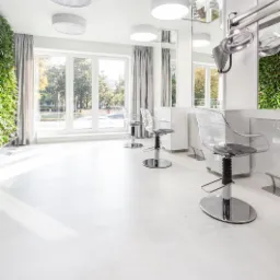 Jasne wnętrze salonu fryzjerskiego z białą posadzką, przezroczystymi krzesłami i zieloną ścianą z roślin.