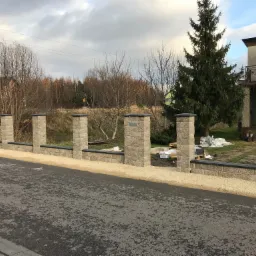 Niski, modułowy płot z szarego kamienia łupanego z czarnymi nakładkami, oddzielający posesję od asfaltowej drogi, z widocznymi materiałami budowlanymi na trawniku za płotem.