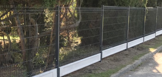 Panelowe ogrodzenie z siatki z podmurówką w kolorze białym, widoczne drzewa w tle.
