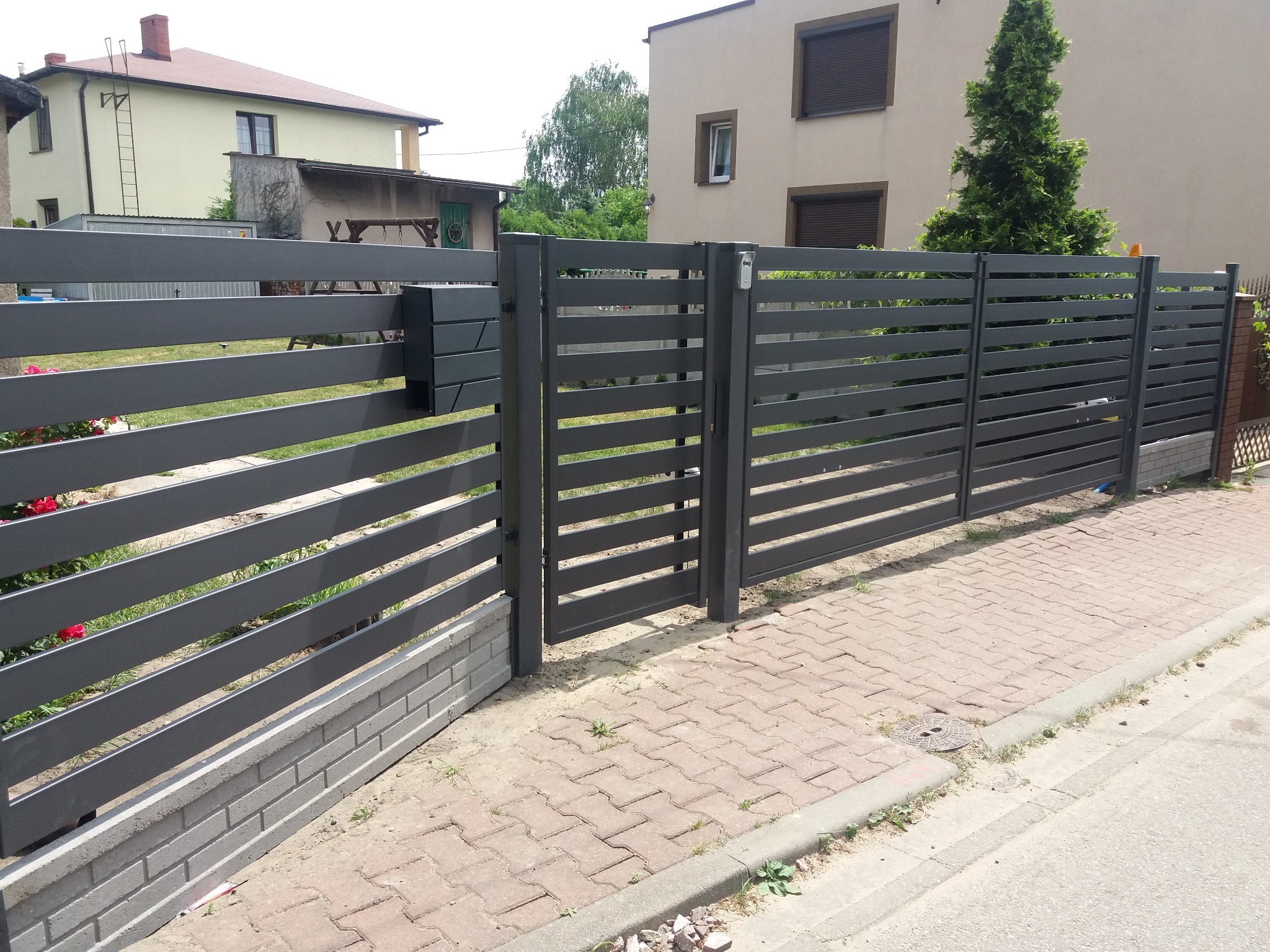 Nowoczesne, poziome ogrodzenie panelowe w kolorze grafitowym z furtką i skrzynką na listy, osadzone na betonowej podmurówce z cegły klinkierowej, widok z zewnątrz na posesję.