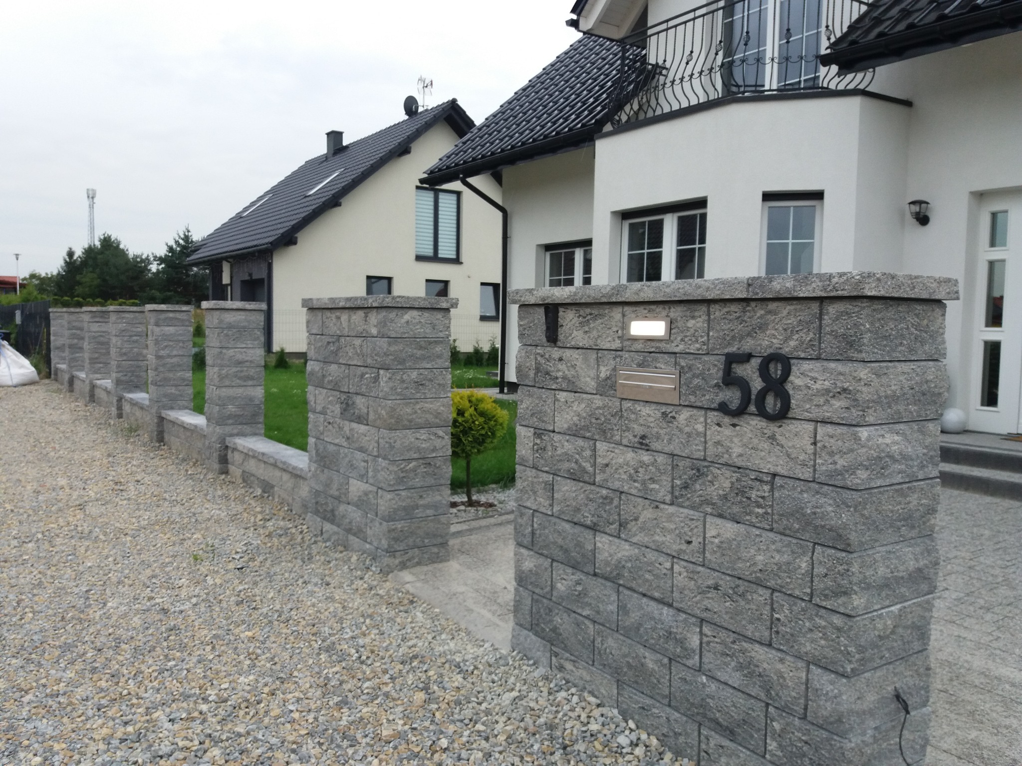 Nowoczesne ogrodzenie z szarej kostki łupanej z numerem domu 58, oświetleniem i żwirowym podłożem, na tle dwóch domów.